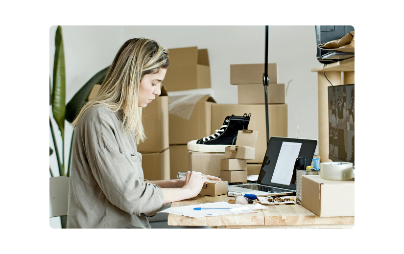 Woman managing eCommerce sales, packing orders with shipping materials, boxes, and a laptop for online business growth.